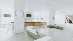 A spacious, modern hotel lobby with a minimalist design, featuring a central seating area, a reception desk, and a large window providing natural light.