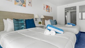 Habitación de hotel con dos camas, ropa de cama blanca, almohadas azules y blancas, y obras de arte en las paredes.
