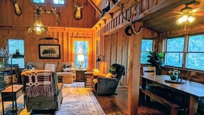 L’intérieur chaleureux d’un chalet avec des murs en bois, une cheminée, un coin repas et une grande fenêtre donnant sur le paysage extérieur.
