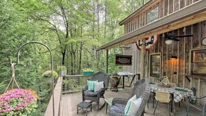 Une terrasse en bois avec des meubles en osier, une table recouverte d’une nappe à carreaux et un pot de fleurs suspendu.