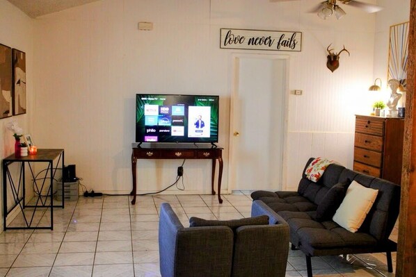 Una sala de estar con un televisor sobre un mueble de madera, un sofá, una pequeña mesa con una planta y un letrero en la pared.
