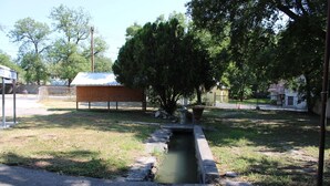 A small outdoor area with a concrete drainage channel, a tree, and a building with a metal roof in the background. - Historic Cozy Cabin Downtown Del Rio (Del Rio)