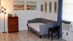 A bedroom with a grey sofa bed, a wooden dresser, a floor lamp, and framed artwork on the wall. - Historic Cozy Cabin Downtown Del Rio (Del Rio)