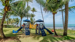 Ein Spielplatz mit Rutschen und Klettergerüsten, umgeben von Palmen und mit Blick auf das Meer.