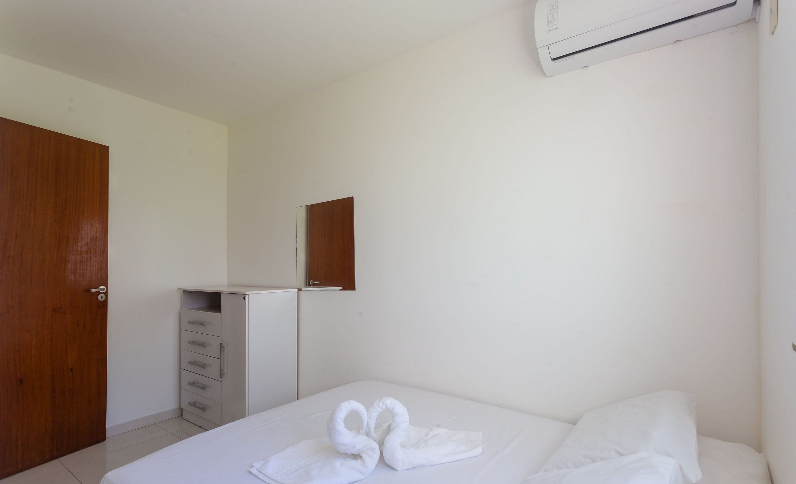 Una camera d'albergo con un letto, una porta in legno, una cassettiera bianca e un condizionatore d'aria a parete.