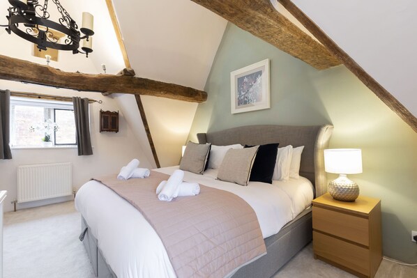 A bedroom with a large bed, a nightstand with a lamp, a window with curtains, and a wooden ceiling beam.