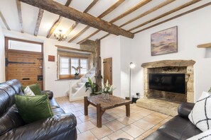 A cozy living room with a fireplace, leather sofas, wooden beams, and a staircase leading to an upper floor. - Charming 3-Bed Cottage in Central Stroud (Gloucestershire)