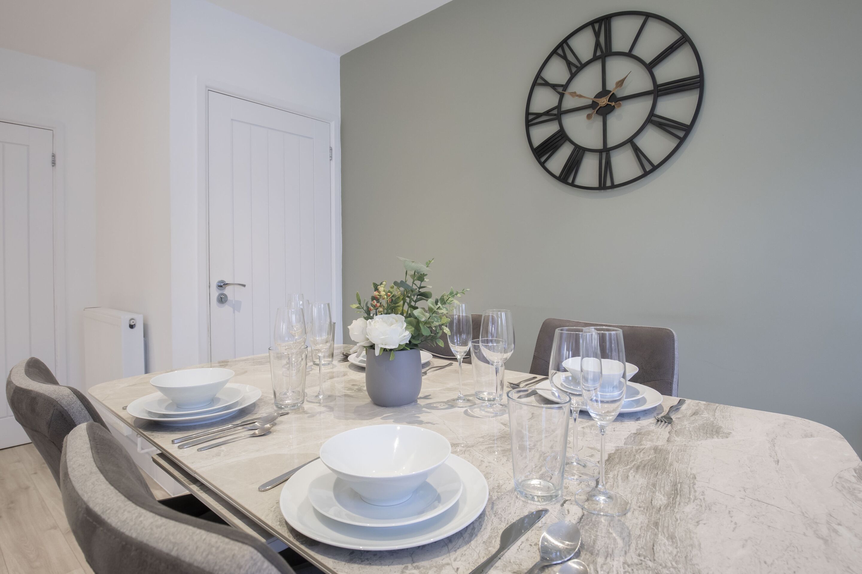 A dining area with a marble table set for four, a large wall clock, and a potted plant.