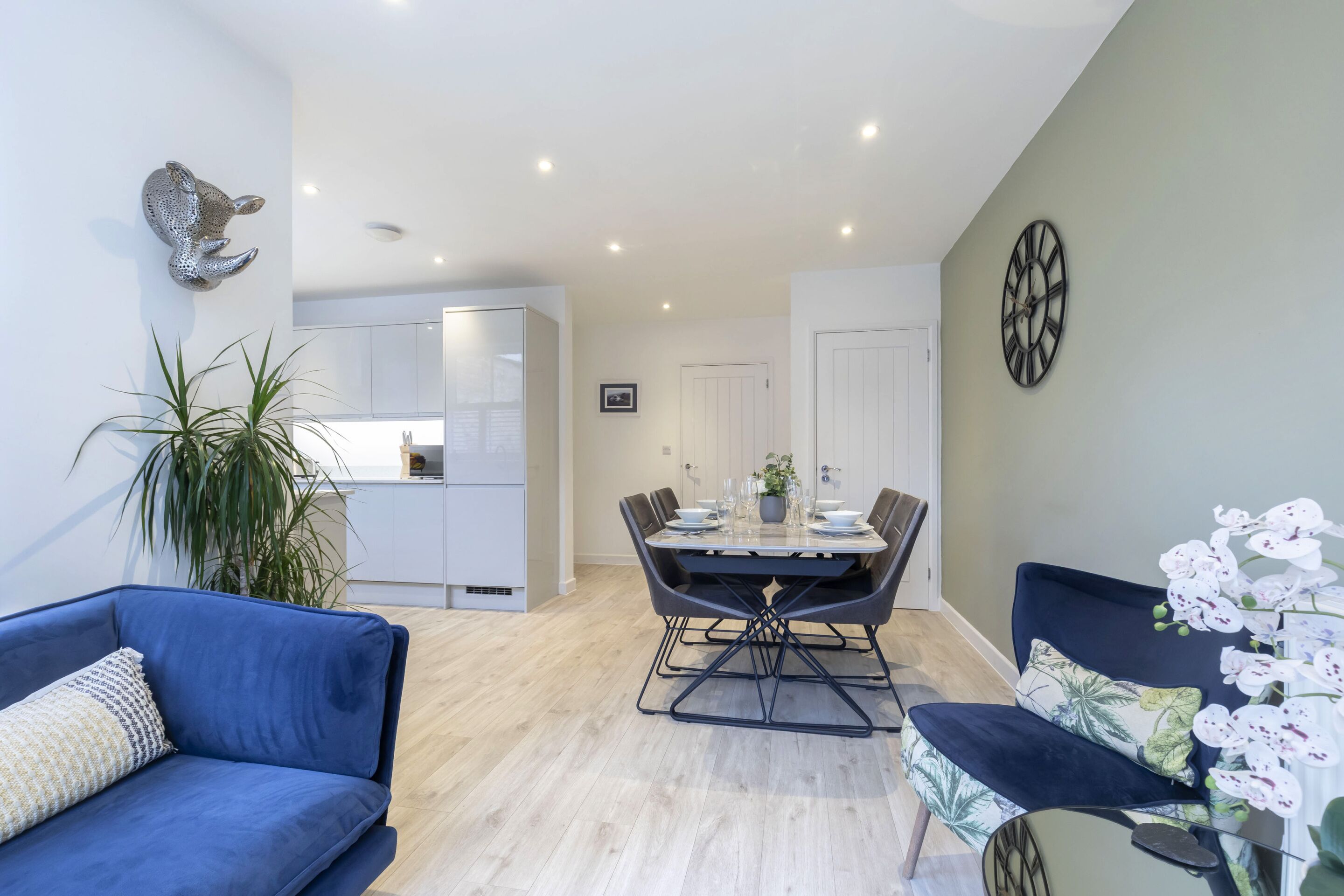 A modern living room with a blue sofa, a dining table set with chairs, a decorative wall clock, and a potted plant.