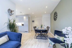 A modern living room with a blue sofa, a dining table set with chairs, a decorative wall clock, and a potted plant.