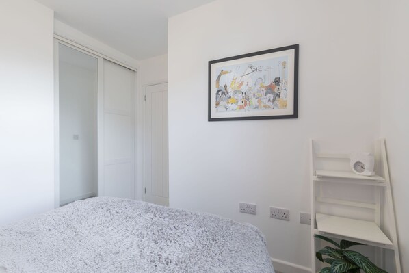 Un dormitorio con cama, una puerta blanca, un cuadro colgado en la pared y una estantería blanca con una planta.