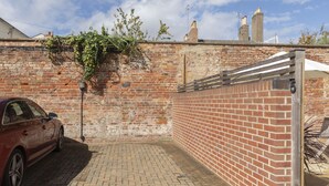 A brick wall with a metal gate, a red car parked on a brick pavement, and a brick building in the background. - Stylish 2-Bed Home in Fairview with Free Parking (Gloucestershire)