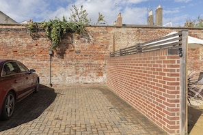 A brick wall with a metal gate, a red car parked on a brick pavement, and a brick building in the background. - Stylish 2-Bed Home in Fairview with Free Parking (Gloucestershire)