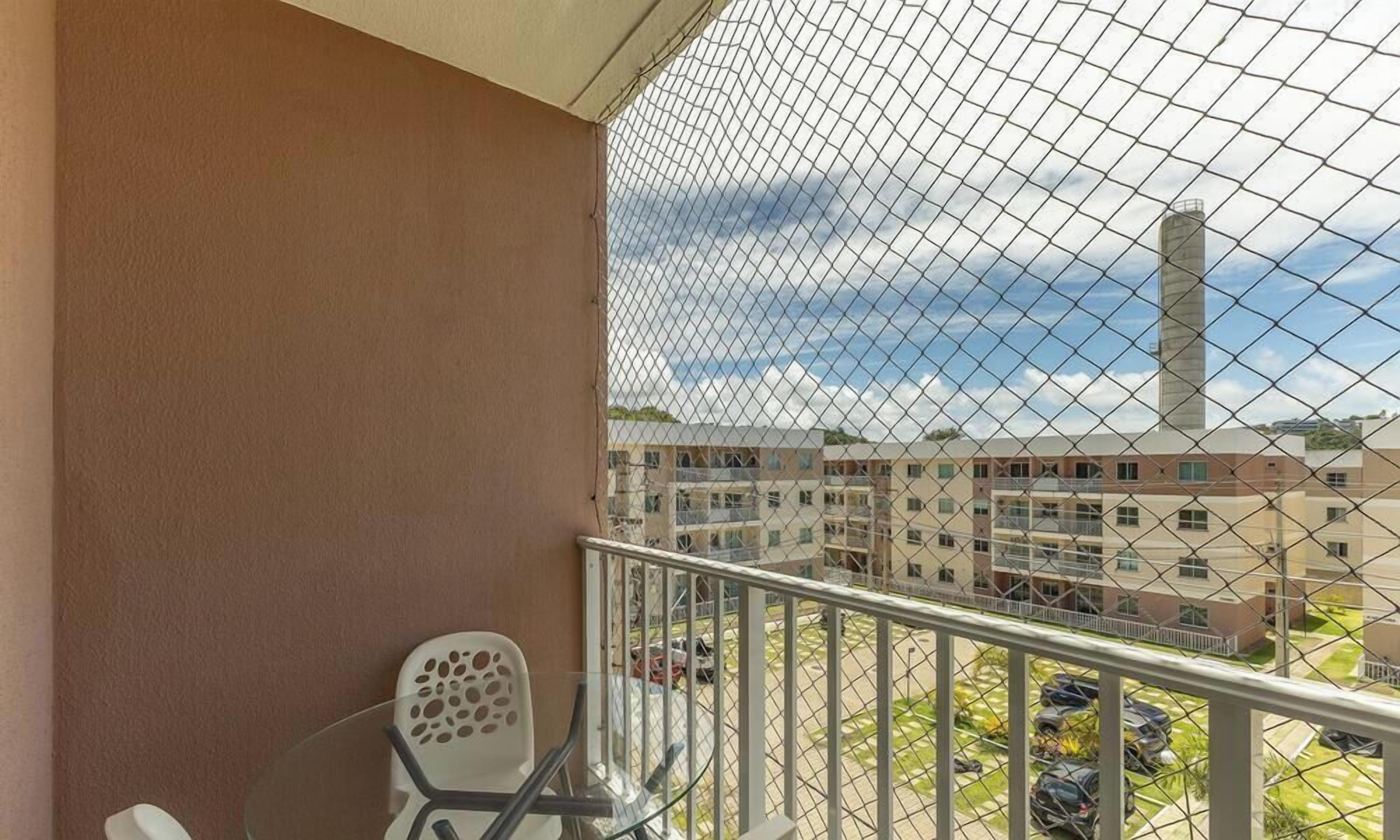 A balcony with a chain-link fence, a white chair, and a view of a multi-story building with a tall chimney.