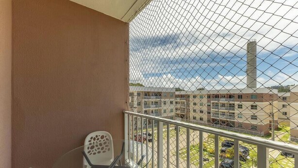 A balcony with a chain-link fence, a white chair, and a view of a multi-story building with a tall chimney. - Apart 450m From the Beach in Cond. w/ Pool Var0302 (Ilhéus)