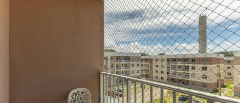 A balcony with a chain-link fence, a white chair, and a view of a multi-story building with a tall chimney.