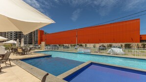 A swimming pool area with multiple pools, outdoor seating, and a large red building in the background. - Apart 450m From the Beach in Cond. w/ Pool Var0302 (Ilhéus)