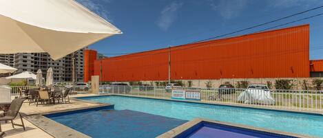 A swimming pool area with multiple pools, outdoor seating, and a large red building in the background.