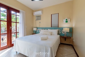 Ein Hotelzimmer mit Bett, Fenster mit Vorhängen, Klimaanlage, Wanduhr und kleinem Tisch mit Lampe.