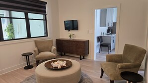 A modern living room with a round ottoman, two armchairs, a small side table, and a wooden cabinet. A flat-screen TV is mounted on the wall, and a kitchen area is visible through an open doorway.