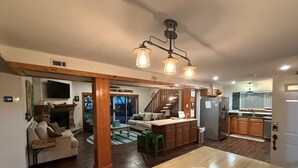 A spacious living area with a wooden dining table, a sofa, and a fireplace. A kitchen with wooden cabinets and a refrigerator is visible in the background.
