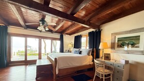 A bedroom with a wooden ceiling, a bed, a desk, and a chair. There is a window with curtains and a view of the outdoors.
