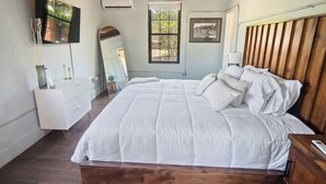 A bedroom with a large bed, a wooden headboard, a white dresser, a mirror, and a window with a view of trees.