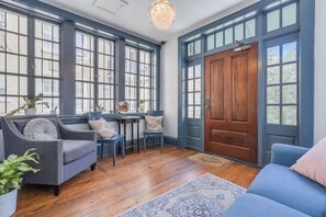 Une pièce dotée de grandes fenêtres, d’une porte en bois, d’un canapé bleu, d’un fauteuil gris et d’une petite table avec des chaises.