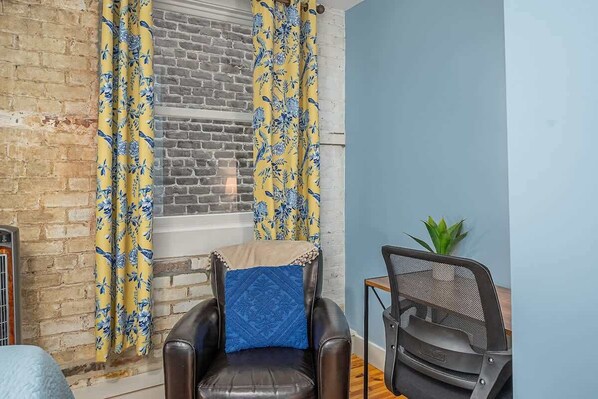Una habitación con pared de ladrillo, ventana con cortinas amarillas, sillón de cuero con cojín azul y una silla de oficina de malla.