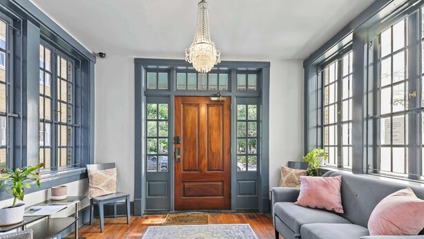 A room with a wooden door, large windows, a chandelier, a grey sofa, and a patterned rug.