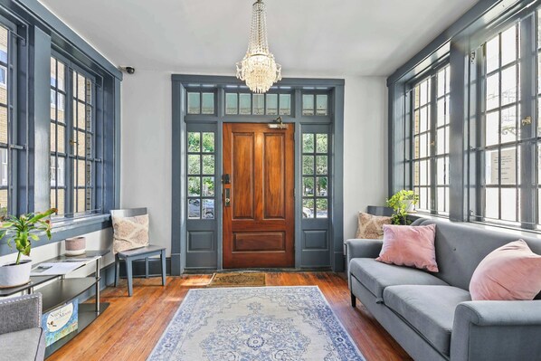 A room with a wooden door, large windows, a chandelier, a grey sofa, and a patterned rug.
