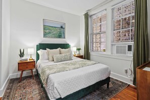 A bedroom with a bed, nightstands, a painting, a window with curtains, and a patterned rug. - Darling Downtown Renovated Historic Condo | Walkable (Savannah)
