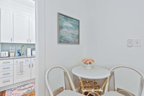Una cocina con armarios blancos, una pequeña mesa de comedor con sillas de mimbre y un cuadro en la pared.