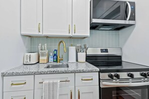 A kitchen with white cabinets, a granite countertop, a toaster, a sink, a microwave, and a stove. - Darling Downtown Renovated Historic Condo | Walkable (Savannah)