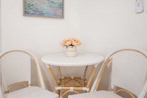 A corner room with a white wicker chair, a small round table with a vase of flowers, and a painting on the wall. - Darling Downtown Renovated Historic Condo | Walkable (Savannah)