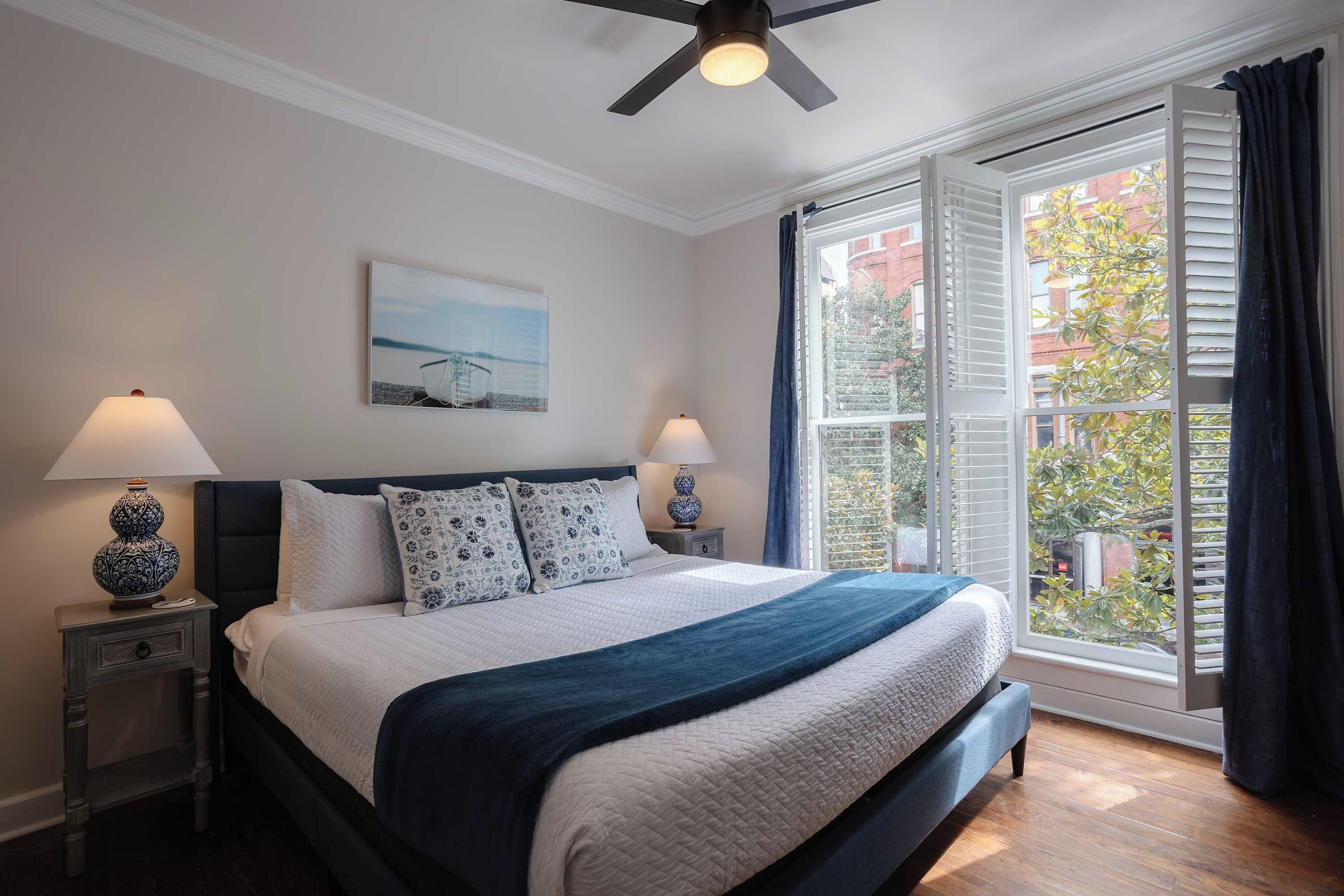 A bedroom with a bed, two bedside tables, a ceiling fan, a window with curtains, and a painting on the wall.