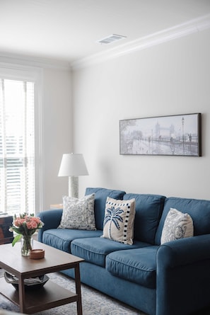 A living room with a blue sofa, decorative pillows, a framed picture, a table with a vase of flowers, and a lamp.