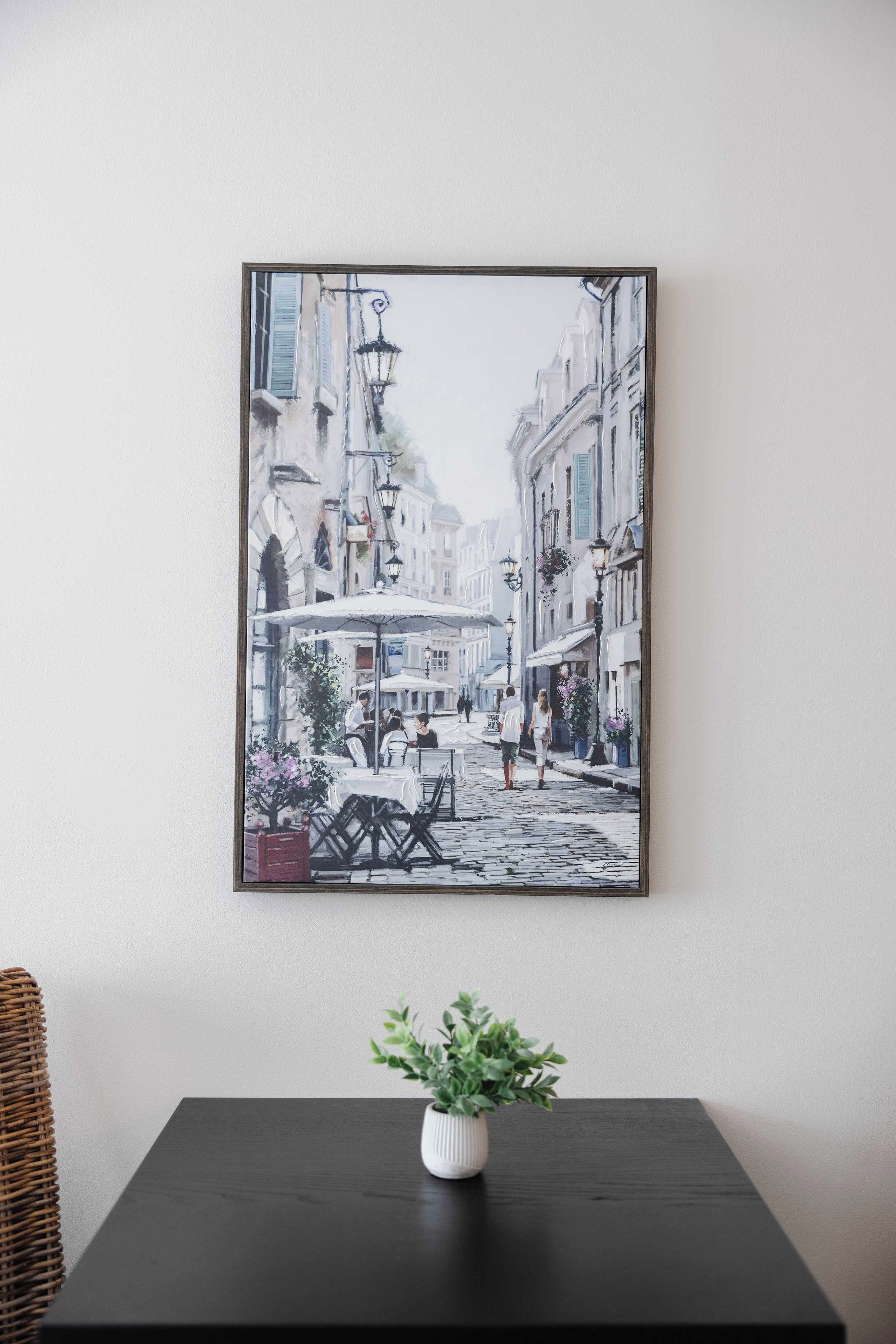 A framed painting of a street scene with buildings, tables, and chairs, mounted on a wall above a table with a potted plant.