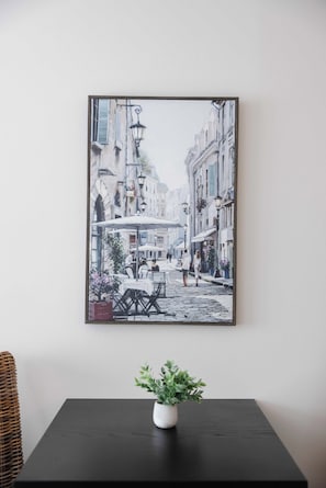 Un tableau encadré représentant une scène de rue avec des bâtiments, des tables et des chaises, accroché au mur au-dessus d’une table sur laquelle se trouve un pot de fleurs.
