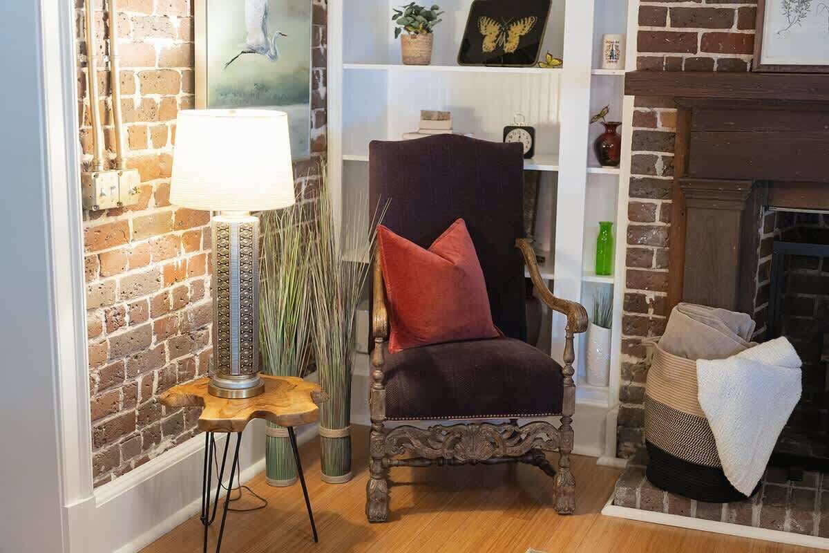 A cozy living room with a brick wall, a fireplace, a dark armchair with a red cushion, a side table with a lamp, and a bookshelf with decorative items.
