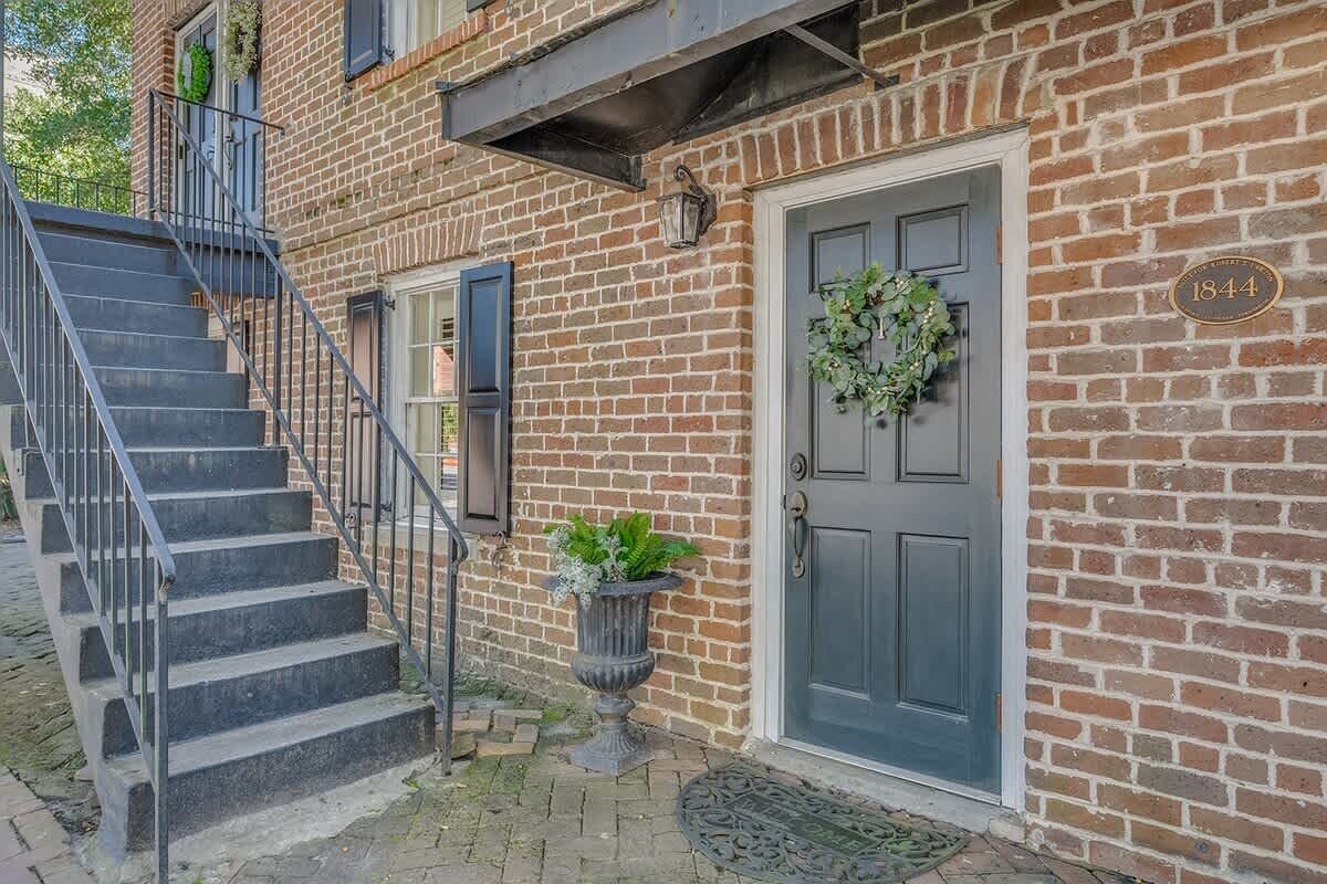 A brick building with a gray door, a number plaque, a staircase, and a potted plant.