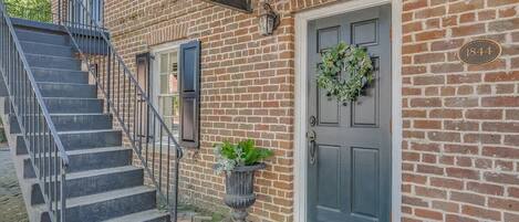 A brick building with a gray door, a number plaque, a staircase, and a potted plant.