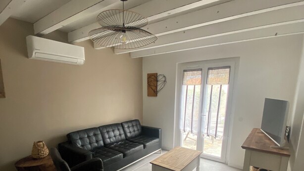Ein Wohnzimmer mit einem schwarzen Ledersofa, zwei schwarzen Sesseln, einem Holztisch, einem Fernseher auf einem Holzschrank und einem Deckenventilator.