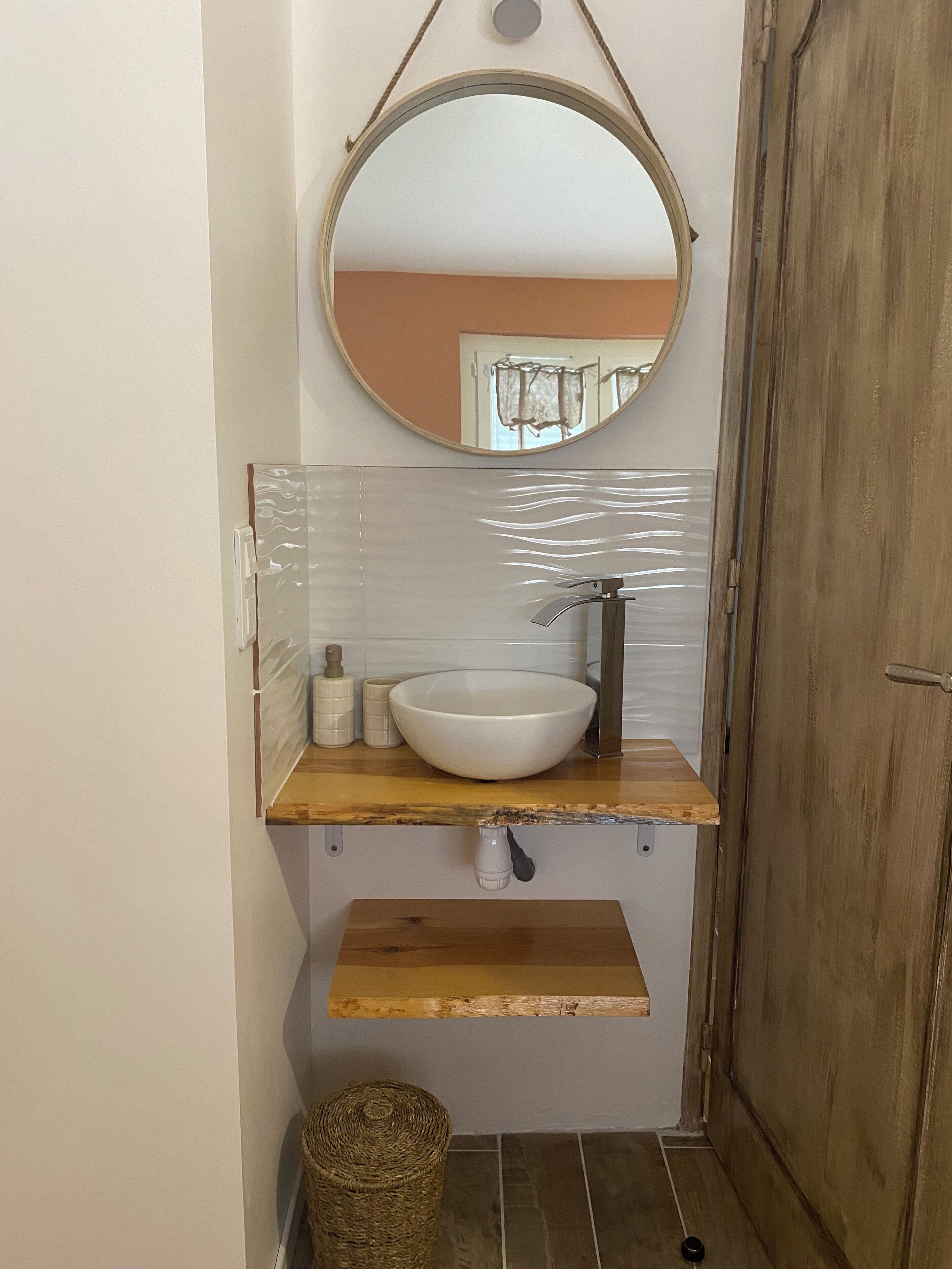 A bathroom with a round mirror, a white sink, and a wooden shelf.