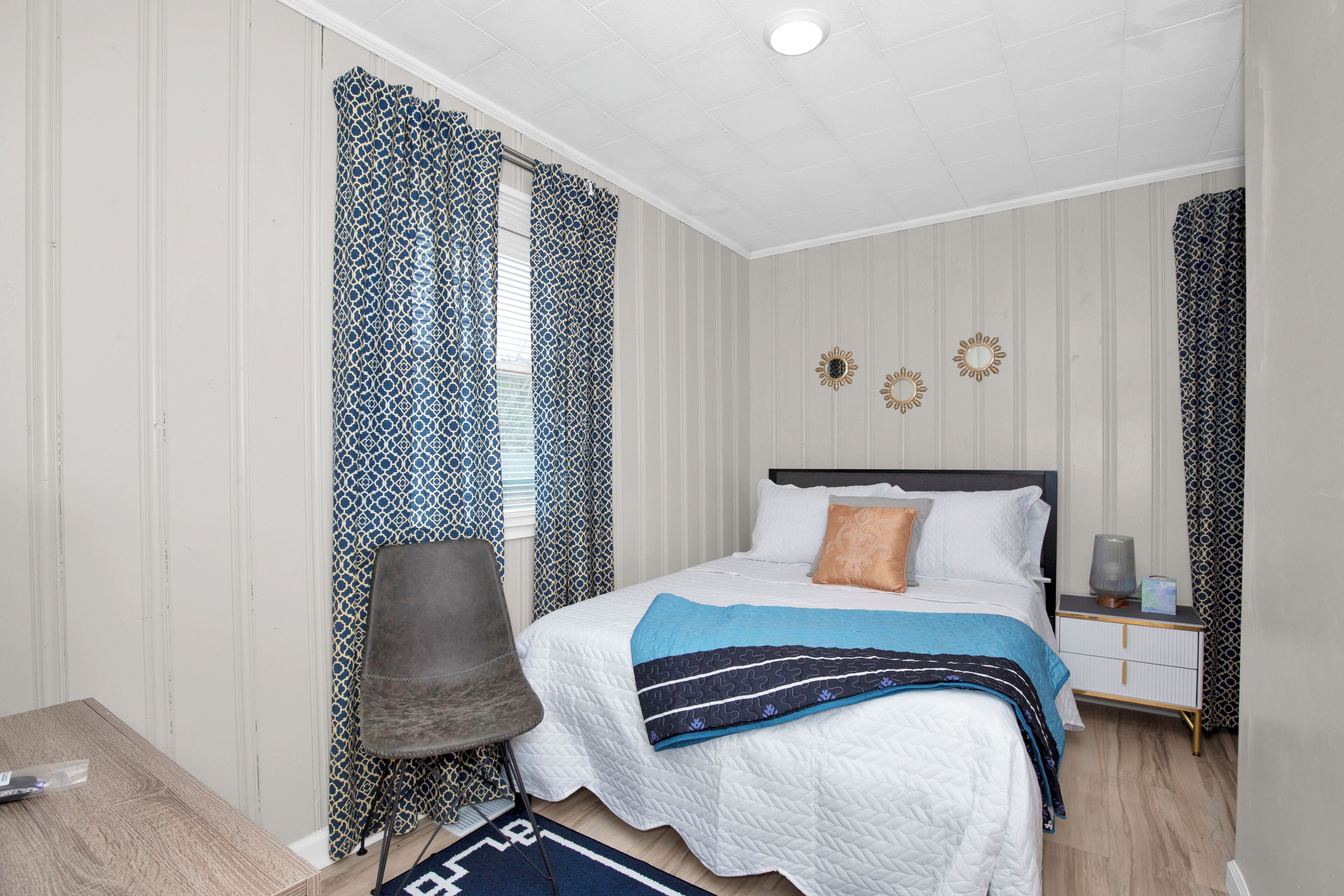 A bedroom with a bed, a chair, a nightstand, and a window with patterned curtains.
