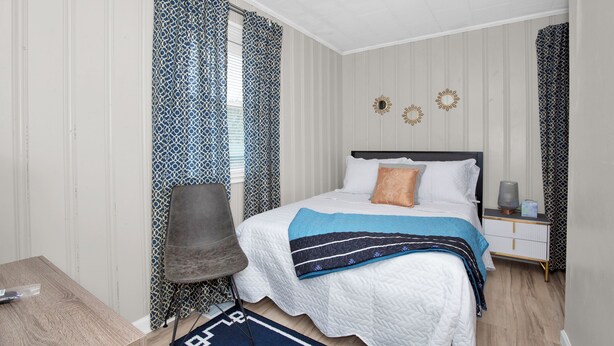 Ein Schlafzimmer mit einem Bett, einem Sessel, einem Nachttisch und einem Fenster mit gemusterten Vorhängen.