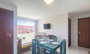 A dining area with a wooden table and chairs, a television mounted on the wall, and a view of the outside through a large window.