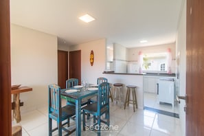 Uma sala de jantar com mesa e cadeiras de madeira, uma cozinha com frigorífico e uma janela com cortinas.