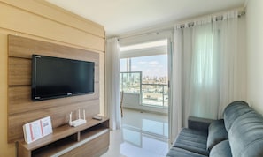 A modern living room with a flat-screen TV mounted on a wooden panel wall, a grey sofa, a small table with a router and remote, and a balcony with a view of a cityscape.