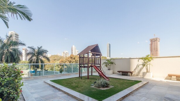 A rooftop garden with a children's play structure, including a slide, surrounded by a low stone wall and palm trees. - Estúdio em Cond no Setor Bueno CEL1804 (Goiânia)